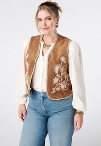 Bruine suède vest met bloemenborduursel aan de voorkant, open ontwerp, gecombineerd met een witte blouse en denim jeans. Stevige textuur, zonder sluitingen.