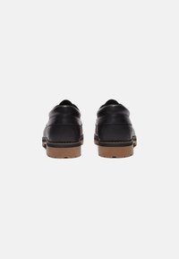 Zapatos de cuero negro con parte trasera redondeada, con detalles de costura y suelas de goma marrón, presentando una textura suave y un diseño minimalista.
