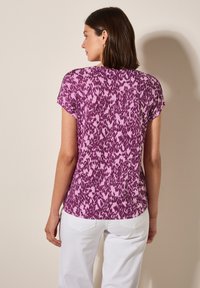Top corto de manga corta con un estampado en tonos morados y rosas, que presenta un corte holgado y una tela suave con un escote redondeado. Combinado con pantalones blancos.