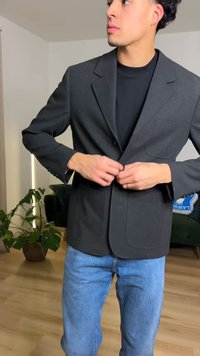 Fekete blézer két első zsebbel, egysoros gombolással és mély kivágású hullámgallérral, kék farmerrel párosítva. Az anyag sima és struktúrált megjelenésű.