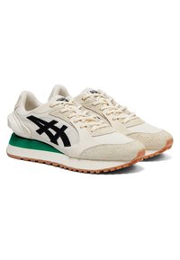 Bílé sportovní boty s texturovaným svrškem, černým logem a zeleným akcentem na patě. Mají zaoblenou špičku a gumovou podešev s protiskluzovým vzorem.