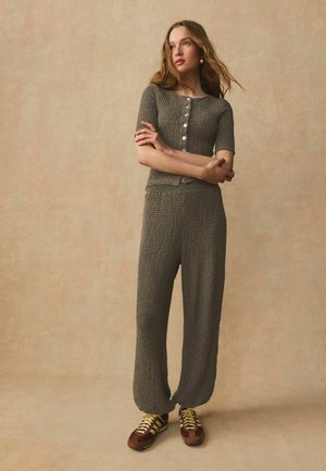 Donna in piedi con le braccia incrociate che indossa un top grigio strutturato con bottoni e pantaloni abbinati a sneakers marroni e gialle su sfondo beige.