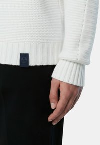 Maglione bianco in maglia con orlo e polsini a coste, con un motivo lavorato a rilievo. Un'etichetta blu navy è visibile in basso a sinistra del maglione.