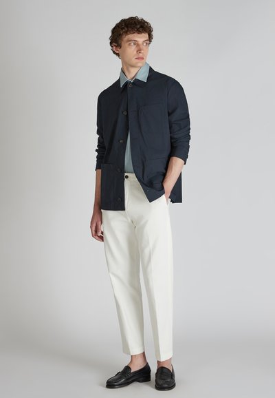 Veste légère navy avec fermetures à boutons, chemise bleu clair, pantalons blancs fuselés et mocassins noirs. Design simple, textures lisses.