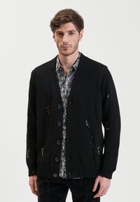 Zwarte gebreide cardigan met een v-hals, voorzien van knopen en versleten details. Draagt over een gestreept shirt.