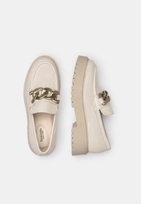 Light beige leather loafers with a chunky sole, featuring a large gold chain accent across the top and visible stitching details.