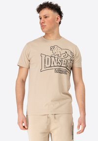 Bež majica od pamuka s kratkim rukavima, na kojoj se nalazi crna grafika lava i tekst "Lonsdale London", okrugli izrez i mekani materijal.