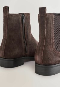 Botas de ante marrón hasta el tobillo con cierre de cremallera, panel lateral de malla, puntera redonda y suela de goma plana. Presenta detalles de costura y un lazo de lengüeta.