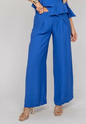Femme portant un pantalon large bleu vif et un haut assorti, avec des sandales à talons hauts dorées et des bracelets argentés aux mains.