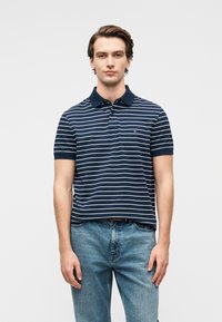 Παντελόνι πόλο σε ναυτικό μπλε χρώμα με λευκές ρίγες, κοντά μανίκια και γιακά. Φοριέται με ανοιχτό μπλε τζιν παντελόνι, με λογότυπο στο στήθος.