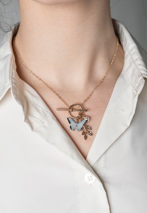 Aukso grandinėlė su mėlynu drugelio pakabuku, apvalia užsegimu ir dekoratyviniais lapų akcentais. Dėvima ant balto apykaklės marškinėlių.