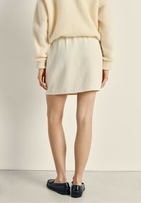 Jupe blanche, de longueur genou, avec une texture lisse et une taille élastique, associée à des mocassins noirs sur des jambes nues, sur un fond neutre.