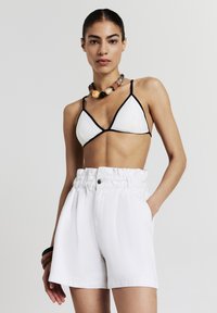 Shorts blancs taille haute avec une finition texturée et une taille froncée. Haut de bikini triangulaire blanc avec des bordures noires. Collier en perles de bois.