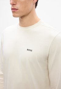 Cămașă bej cu mânecă lungă, fabricată dintr-un material moale, având un decolteu rotund și un mic logo negru "BOSS" pe partea stângă a pieptului.