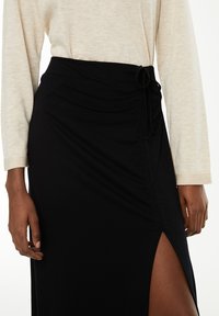 Falda negra con una abertura lateral, presenta un diseño fruncido y un detalle de lazo en la cintura. Fabricada con una tela suave y elástica.