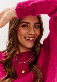 Jaskrawy różowy, puszysty sweter, warstwowe złote i kolorowe koraliki naszyjników, długie falowane brązowe włosy i uśmiechnięta twarz na białym tle.