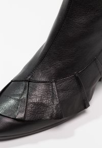 Bottine en cuir noir avec bout pointu et détails plissés sur le côté, texture lisse et design minimaliste.