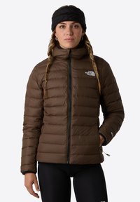 Brązowa kurtka puchowa z pionowymi szwami, wysokim kołnierzem i zapinanymi na zamek kieszeniami. Noszona z czarną opaską na włosy, w połączeniu z czarnymi leginsami.