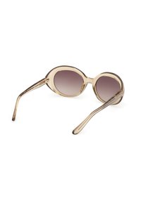 Lunettes de soleil ovales beige transparent avec des verres marron dégradés et des branches courbées, posées sur une surface blanche.