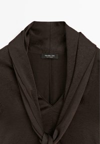 Top in tessuto marrone e strutturato con scollo a V e colletto drappeggiato, caratterizzato da un dettaglio con fiocco frontale. Etichetta con la dicitura "Massimo Dutti, made in Morocco."