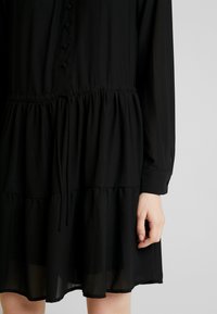 Černé šaty vyrobené z lehkého materiálu, s vrstveným designem, knoflíky v přední části a dlouhými rukávy se staženým pasem.