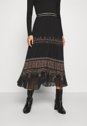 Jupe midi noire plissée avec des motifs cachemire près de l'ourlet, associée à un haut à manches longues et des bottines noires à talons, portée par une personne debout.