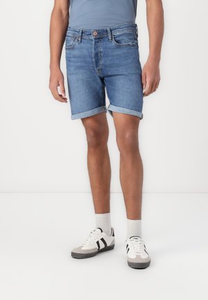 Homme portant un short en jean bleu moyen retroussé, une chemise bleue, des chaussettes blanches et des baskets blanches avec des rayures noires.