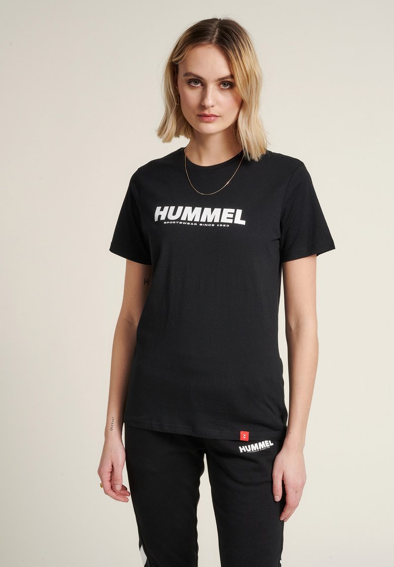 T-shirt en coton noir avec un grand logo blanc "HUMMEL" sur la poitrine. Manches courtes et coupe décontractée. Associé à un pantalon noir.