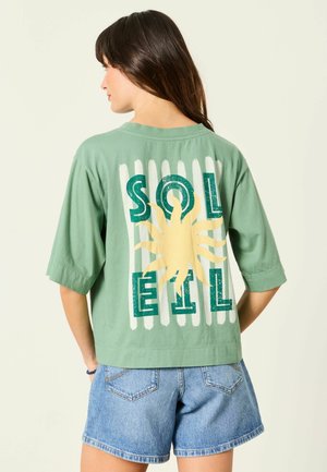 Frau trägt grünes Shirt mit gelber Sonnen-Grafik und dem Schriftzug „SOLEIL“ auf dem Rücken, kombiniert mit blauen Jeansshorts, steht vor einfachem Hintergrund.