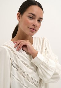 Blouse blanche avec des détails en dentelle et des manches bouffantes, présentant un design en V et des motifs complexes sur le tissu. Texture lisse.
