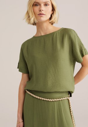 Femme portant un haut à manches courtes texturé vert olive et une jupe avec une ceinture chaîne dorée et des boucles d'oreilles créoles dorées sur un fond neutre.