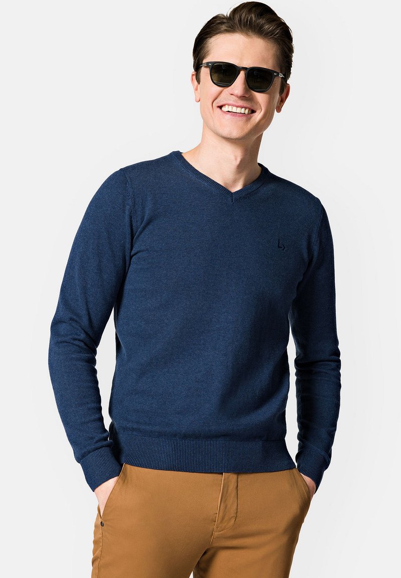 Granatowy sweter dzianinowy z dekoltem w serek, wykonany z miękkiego materiału. Posiada prążkowany dół i mankiety, noszony z khaki spodniami. W zestawie ciemne okulary przeciwsłoneczne.