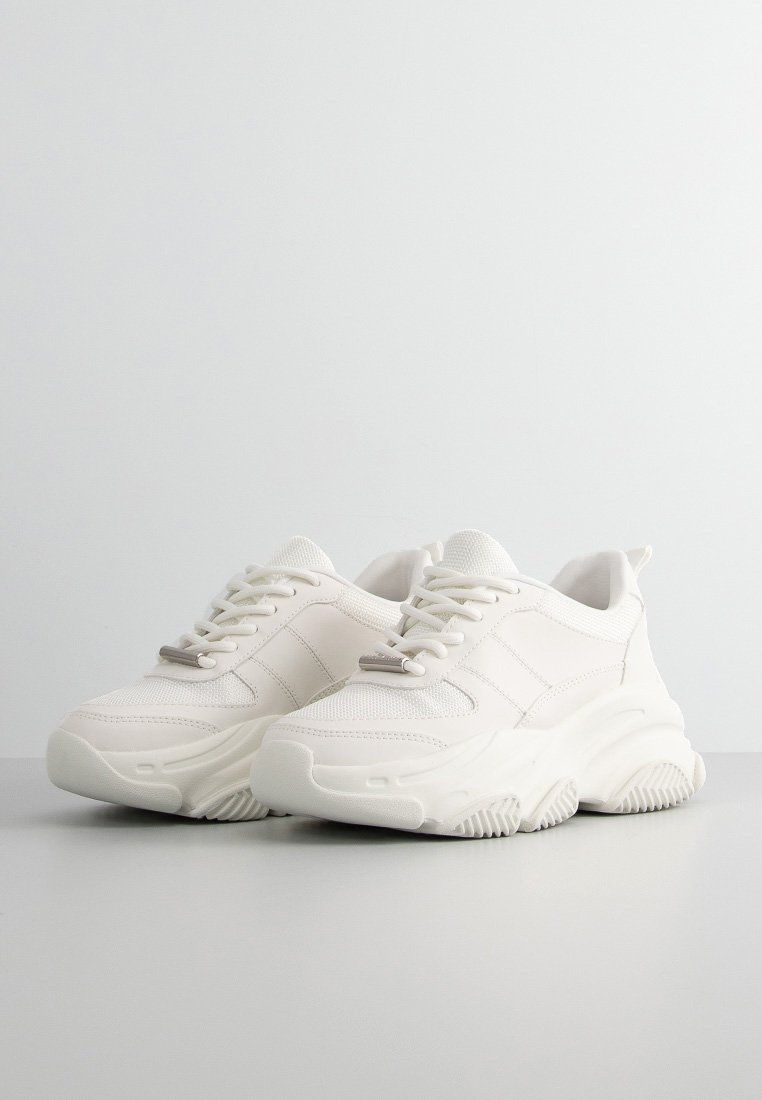 Zapatillas blancas con suela gruesa, parte superior de malla y sintético, punta redonda, cordones planos y acentos laterales. Suela texturizada para tracción.