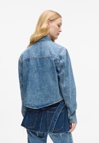 Τζάκετ από τζην σε ανοιχτό μπλε χρώμα, με υφή, μακριά μανίκια και μια ξεχωριστή σχεδίαση σε επίπεδα στην πλάτη με δύο πίσω τσέπες.