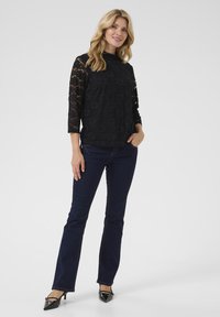 Top lungo in pizzo nero con maniche lunghe e motivo floreale, abbinato a jeans a palazzo blu scuro e décolleté nere a punta, in piedi contro uno sfondo grigio.