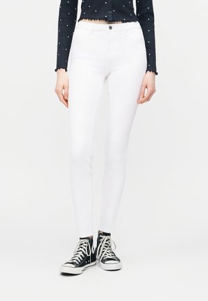 Γυναίκα που φοράει λευκό στενό παντελόνι, μαύρο μακρυμάνικο μπλουζάκι με λευκές βούλες και μαύρα ψηλά αθλητικά παπούτσια με λευκά κορδόνια, στέκεται σε λευκό πάτωμα.