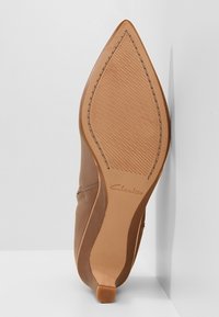 Hnědý kožený podpatek s špičatou špičkou, hladkou texturou a gumovou podrážkou s vzorem rybí kosti. Logo "Clarks" je embosováno na stélce.