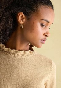 Beige gerippter Pullover mit einem gerafften Ausschnitt. Nahaufnahme eines Profils, das Ohrringe und strukturiertes Haar vor einem neutralen Hintergrund hervorhebt.