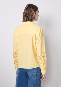 Una donna con i capelli corti castani indossa una blusa gialla a maniche lunghe con design traforato sulle maniche e jeans blu, in piedi contro un muro neutro.