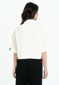 White cropped blouse with short sleeves, featuring a straight cut and subtle back seam, paired with black trousers. Smooth, lightweight fabric.