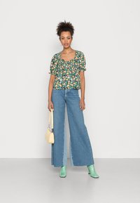 Blusa de manga curta com estampado floral em verde escuro, combinada com jeans de ganga de cintura alta e perna larga. Acessórios incluem uma mala amarela e botas verdes.