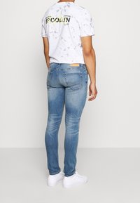 Herrens ljusblå slim-fit jeans med blekta detaljer, kombinerade med en vit t-shirt med grafisk tryck och svarta bokstäver. Vita sneakers.