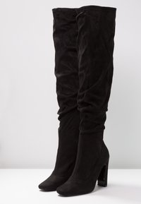 Botas de ante negras por encima de la rodilla con un diseño arrugado y punta afilada, que cuentan con un alto tacón cuadrado y un detalle de cremallera lateral.