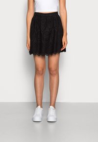 Zwarte, knie-lange rok met oogjesdetails, gegolfde zoom en elastische tailleband. Gecombineerd met witte sneakers voor een casual look.