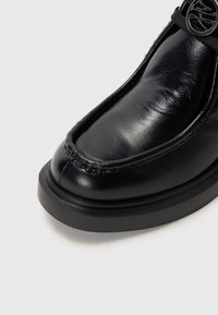 Chaussure en cuir noir avec une finition brillante, bout rond, couture contrastante, et semelle en caoutchouc noir, dotée d'un accent décoratif en métal argenté.