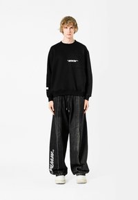 Sudadera negra con un bolsillo frontal y logo blanco, combinada con pantalones de mezclilla negros de corte holgado que presentan un detalle en texto blanco. Zapatillas blancas.