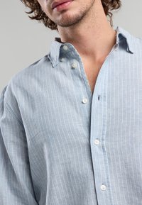 Uomo che indossa una camicia a bottoni leggera di colore azzurro, con sottili righe bianche, parzialmente sbottonata, che mostra il collo e una leggera peluria sul petto.