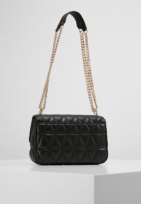 Musta tikattu käsilaukku, jossa on geometrinen kuvio, kultaketjuhihna ja teksturoitu nahka-pinta.