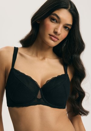 Młoda kobieta z długimi ciemnymi włosami, ubrana w czarny koronkowy biustonosz z fiszbinami, pozująca na jasnym, neutralnym tle.