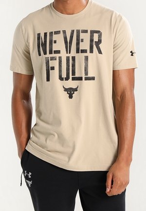 Homme portant un t-shirt beige avec le texte "NEVER FULL" et un logo de taureau, associé à un pantalon noir arborant les logos Under Armour.